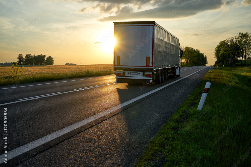 Truck departing towards the horizon on an asphalt road in a rural countryside at sunset.