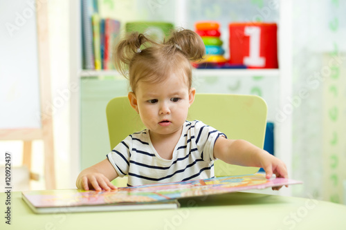 little smart girl looking at book while sitting on chair in nursery