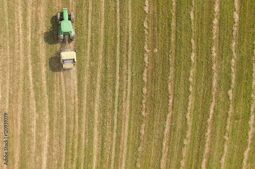 Foto Aerial view of tractor with round baler rolling bales of straw on harvested fiel