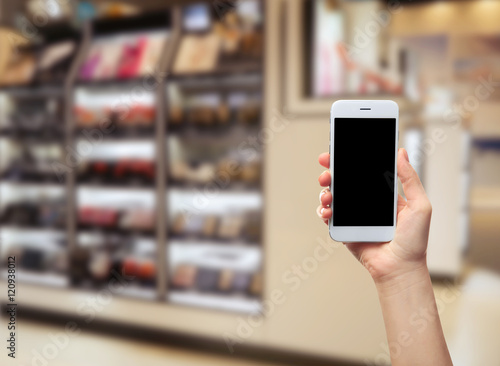 Female hand holding smartphone on blurred cosmetics shelves background.