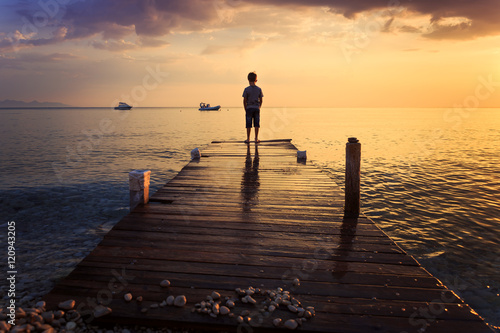 Child standing on a wooden pier at bright dramatic sunrise and looking at the sea