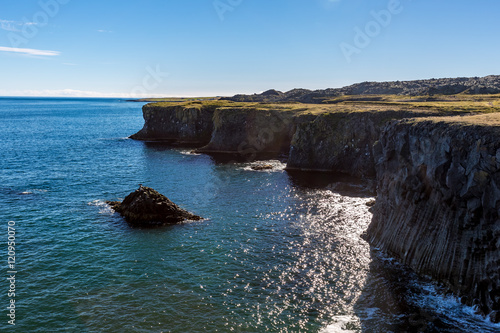 Beautiful Icelandic coast with high volcanic cliffs, the blue ocean in a sunny day. A black sea bird is spreading its wings on a small rock in the water.