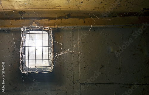  Shining old light covered in metal grill and cobwebs against dirty, peeling painted wall.