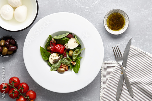 Italian salad with fresh basil leaves, olives, capers, walnuts, mozzarella cheese and red pepper on stone background.