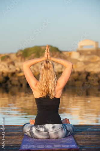Smiling athletic female seated on dock with hands overhead in Mountain Pose. 