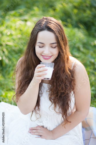 Happy attractive girl sitting on grass and drinking milk in summer outdoors. Close up portrait. Healthy lifestyle. Breakfast in park.