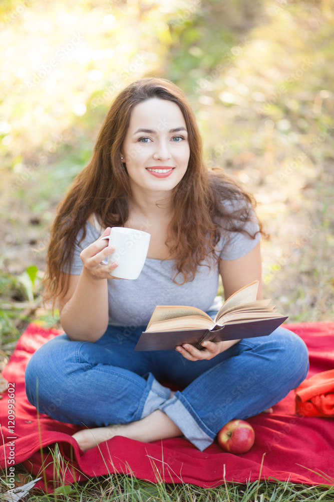 Happy smiling attractive girl sitting on wrap reading book and drinking tea in autumn outdoors. Education and student concept. Study in park Happy smiling attractive girl sitting on wrap reading book and drinking tea in autumn outdoors. Education and student concept. Study in park