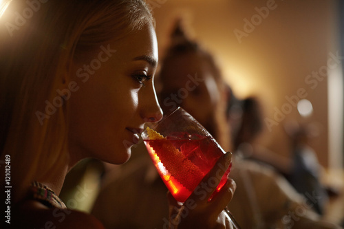 People, leisure and lifestyle concept. Beautiful coquettish girl with fair hair holding glass of ice drink, celebrating her birthday at night club. Pretty female with flirting smile drinking cocktail