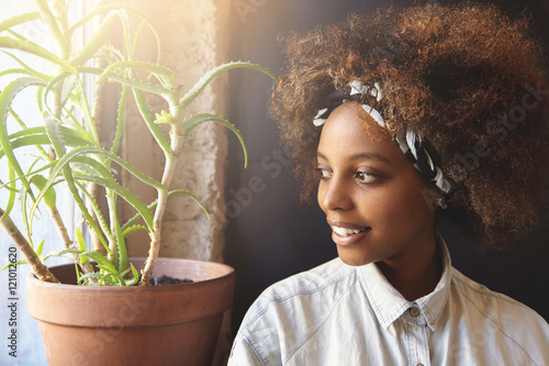 Headshot of young beautiful hipster woman with Afro hairstyle wearing do-rag on her head, looking outside through window with joyful smile, contemplating sunrise. Black girl spending day-off at home photo