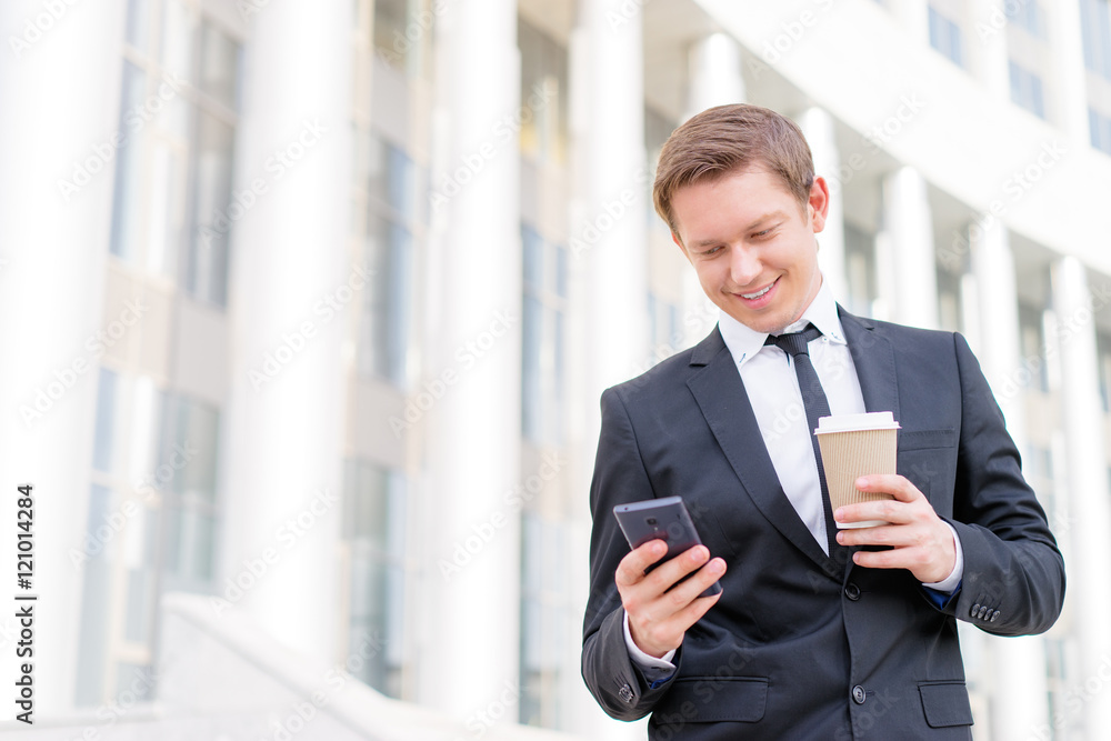 Work and coffee break. Cheerful young businessman using smartphone and holding cup of beverage. Work and coffee break. Cheerful young businessman using smartphone and holding cup of beverage.