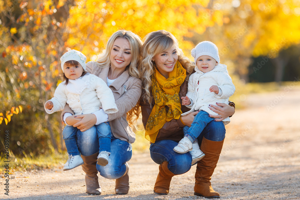 Fototapeta premium Two young mothers,a blonde,dressed in blue jeans and leather jackets,walk in the Park day on the background of beautiful yellow autumn nature with young daughters,dressed in white jackets