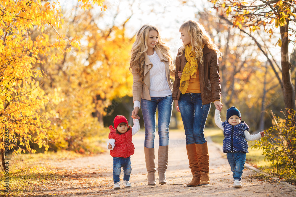 Two young mothers,a blonde,dressed in blue jeans and leather jackets,walk in the Park day on the background of beautiful yellow autumn nature with young daughters,dressed in red and blue jackets Two young mothers,a blonde,dressed in blue jeans and leather jackets,walk in the Park day on the background of beautiful yellow autumn nature with young daughters,dressed in red and blue jackets