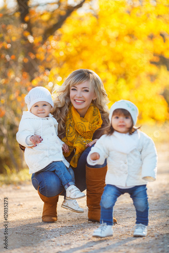Young mother with long curly blond hair and blue eyes,dressed in blue jeans and brown shoes walking in yellow autumn Park with two young girls,dressed in blue jeans,a white jacket and white cap