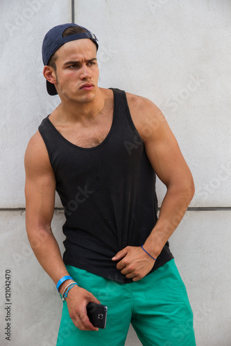 Young, caucasian, strong and handsome man wearing a black tank top and a cap. He has the smart phone in his hand. He looks with a seductive face while resting his arm on the wall.