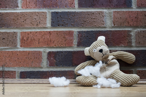 A sad, abandoned teddy bear with stuffing and filling falling from a hole in his ripped and torn tummy and sitting alone against a brick wall. photo