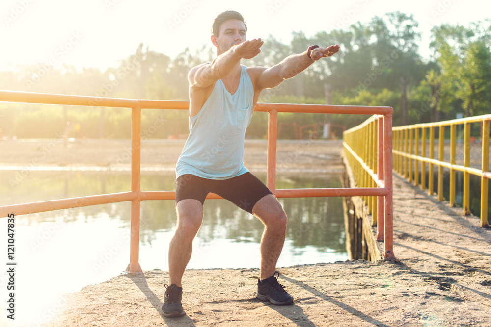 Fototapeta premium Fit man warming up doing squats stretching arms forward outdoors.