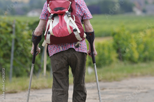eine gehbehinderte seniorin geht an krücken mit rucksack einen weg entlang