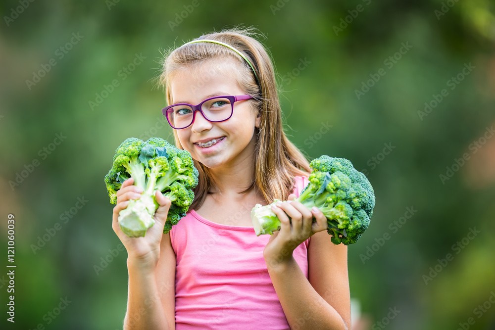 Funny cute girl holding in hands red cabbage and broccoli. Blurred background in garden. Pre-teen young girl with glasses and teeth braces. Funny cute girl holding in hands red cabbage and broccoli. Blurred background in garden. Pre-teen young girl with glasses and teeth braces.
