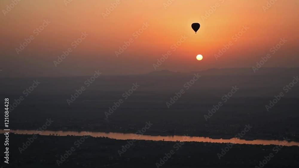 Silhouette of hot air balloon flying at dawn over rural farmland landscape with river and sunrise background Silhouette of hot air balloon flying at dawn over rural farmland landscape with river and sunrise background
