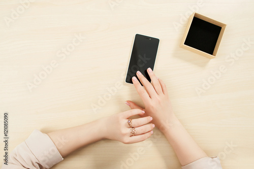 Woman using smartphone, mockup, user pov. Top view on female hands on wooden table touching phone screen and little carton box, free space