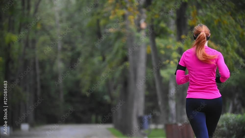 Runner young woman running in autumn park exercising outdoors at rain, pink training suit, rear view, gadget on the hand, slow motion Runner young woman running in autumn park exercising outdoors at rain, pink training suit, rear view, gadget on the hand, slow motion