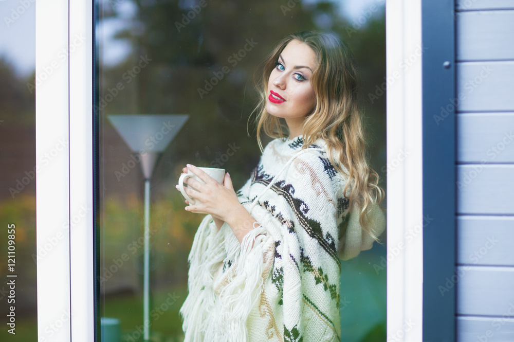 Fototapeta premium Young beautiful blonde woman with cup of coffee standing by the window. Garden reflection on the glass. 