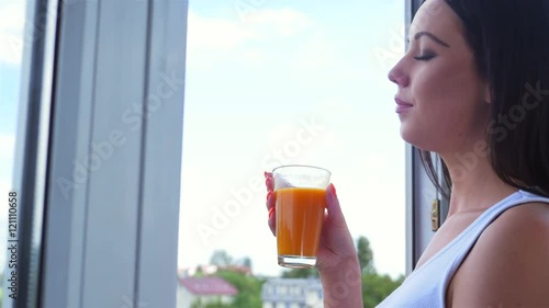 Young woman looking out the window and drinking carrot juice. City background
