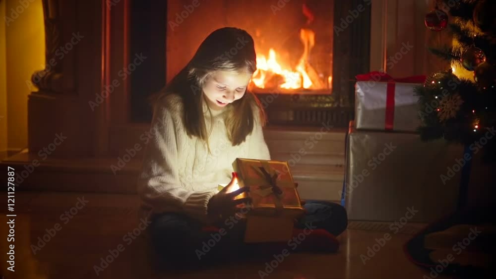 Cute girl sitting in dark living room at burning fireplace and opening box with Christmas present Cute girl sitting in dark living room at burning fireplace and opening box with Christmas present