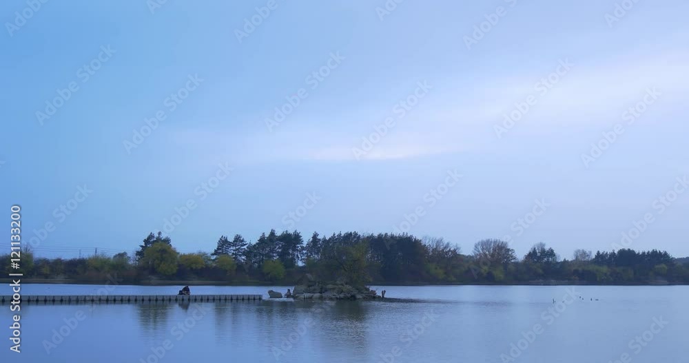 Man Sits Down on Wooden Pier on River Small Island Overgrown With Trees Autumn Landscape Yellow and Green Leaves Dusk Trees on the Opposite Side River Bank Man Sits Down on Wooden Pier on River Small Island Overgrown With Trees Autumn Landscape Yellow and Green Leaves Dusk Trees on the Opposite Side River Bank