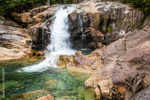 Young man and woman sitting by the base of the waterfall in the forest