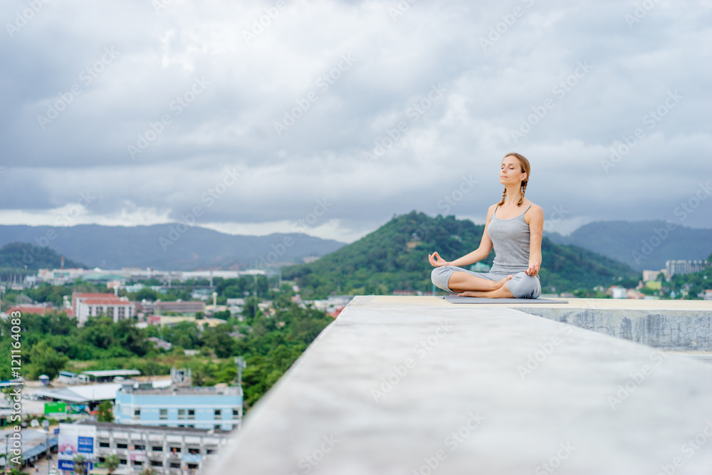 Yoga on rooftop. Happy young woman in lotus pose on roof with city and mountains view.
