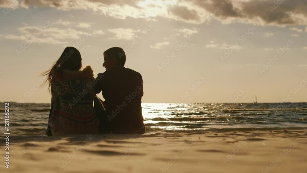 Silhouettes of couples in love, sitting on the beach, watching the sunset and a board with a sail. Back view Silhouettes of couples in love, sitting on the beach, watching the sunset and a board with a sail. Back view