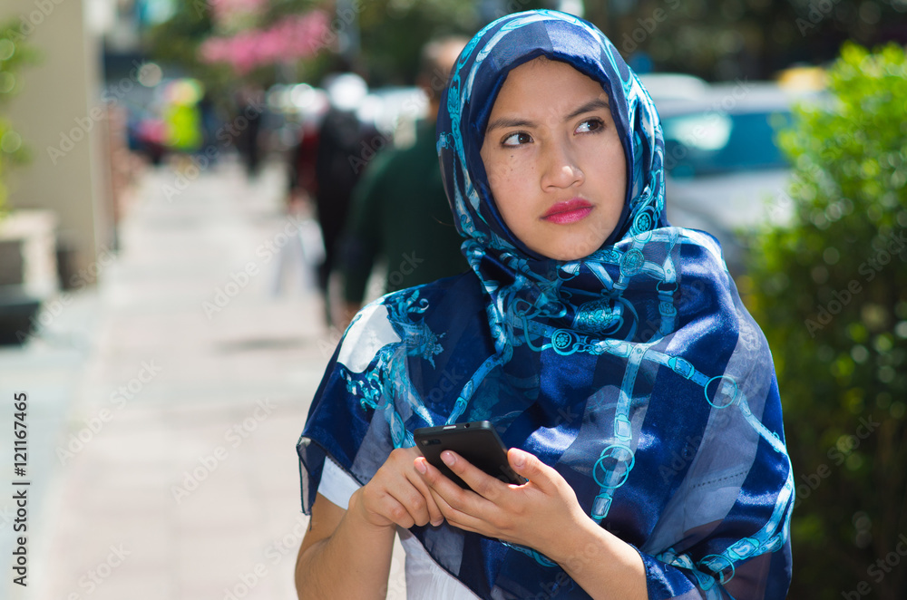 Beautiful young muslim woman wearing blue colored hijab, using mobile phone in outdoors urban background Beautiful young muslim woman wearing blue colored hijab, using mobile phone in outdoors urban background