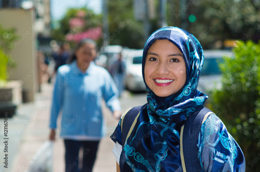 Beautiful young muslim woman wearing blue colored hijab and backpack, posing happily in street smiling to camera, outdoors urban background Beautiful young muslim woman wearing blue colored hijab and backpack, posing happily in street smiling to camera, outdoors urban background