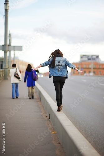 Brave girl in a blue denim jacket goes on the curb, intently trying to keep his balance, on a busy city street.