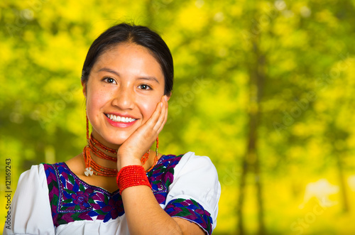 Headshot beautiful young woman wearing traditional andean blouse with red necklace, posing for camera touching face using hands while smiling happily, green forest background