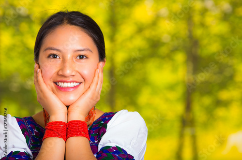 Headshot beautiful young woman wearing traditional andean blouse with red necklace, posing for camera touching face using hands while smiling happily, green forest background