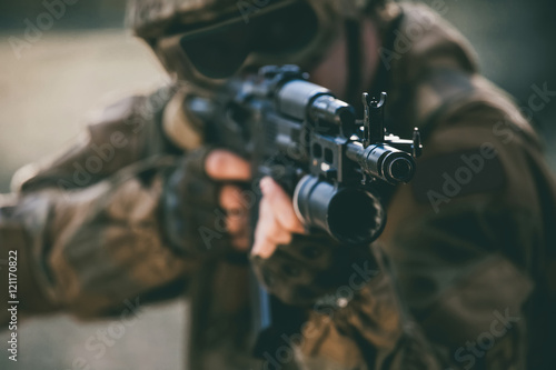 The soldier in the performance of tasks in camouflage protective gloves helmet and tinted glasses holding a machine gun takes aim for shot. War Zone.