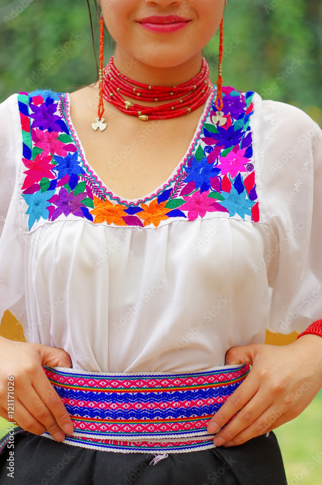 Closeup beautiful hispanic woman wearing black skirt, traditional andean white blouse with colorful decoration around neck and waist, matching red necklace, bracelet, ear ring, garden background