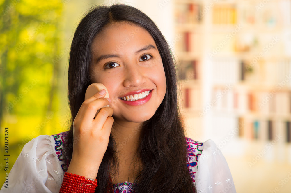 Beautiful hispanic woman wearing white blouse with colorful embroidery, using ponge applying makeup, smiling to camera Beautiful hispanic woman wearing white blouse with colorful embroidery, using ponge applying makeup, smiling to camera
