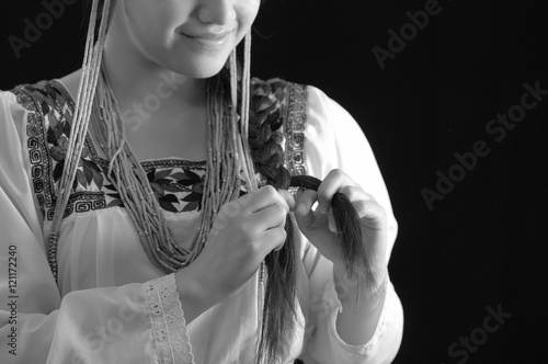 Beautiful young hispanic woman wearing light colored blouse with traditional embroided edges, braiding her own hair while smiling, dark studio background, black and white edition