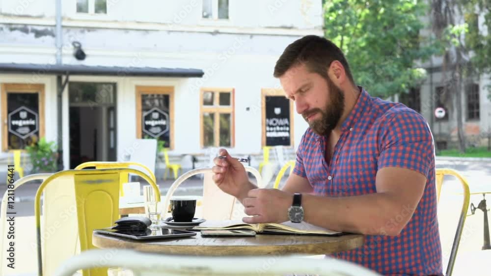 Man makes notes from smartphone to notebook. He is sitting at the table in cafe. He is young and has beard. Man is dressed in checkered shirt. Slider shot, righ
