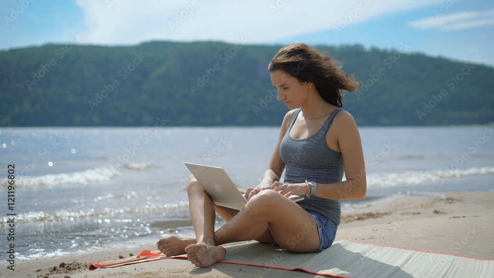 A beautiful sexy young lady is sitting and smiling at a beach using laptop