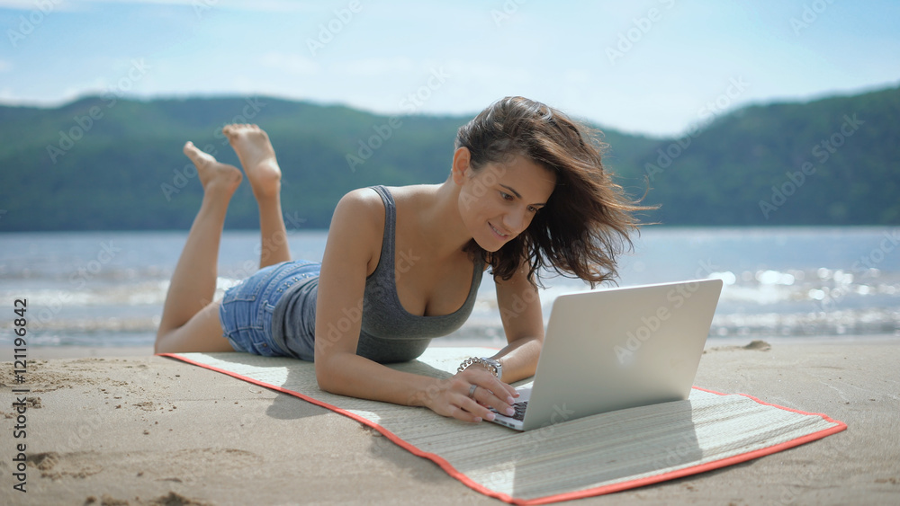 Fototapeta premium A beautiful sexy young smiling lady is laying on her tommy at a beach using laptop