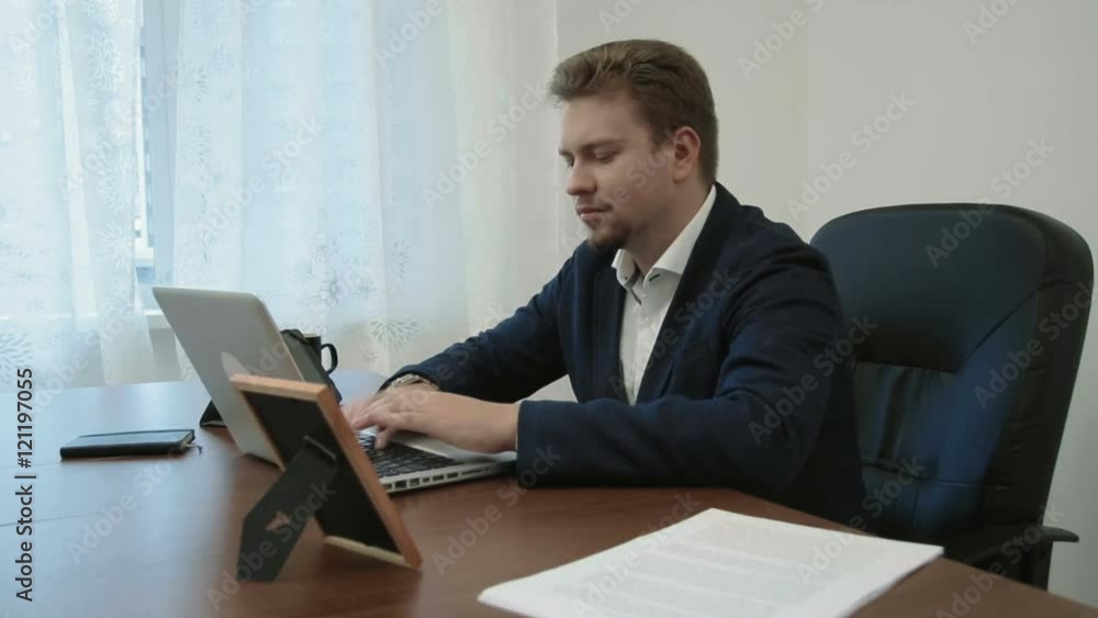 Young businessman working in his office in front of the laptop computer. Then looks at the picture of his family takes it, smiles and continues working Young businessman working in his office in front of the laptop computer. Then looks at the picture of his family takes it, smiles and continues working