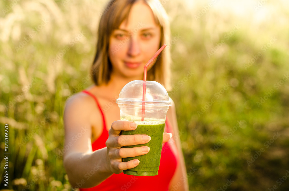 face of a smiling young woman dressed in a red sports shirt useful in the hands green energy and nutritional smoothie cocktail with banana mint lettuce lime cucumber on the background blurred nature face of a smiling young woman dressed in a red sports shirt useful in the hands green energy and nutritional smoothie cocktail with banana mint lettuce lime cucumber on the background blurred nature