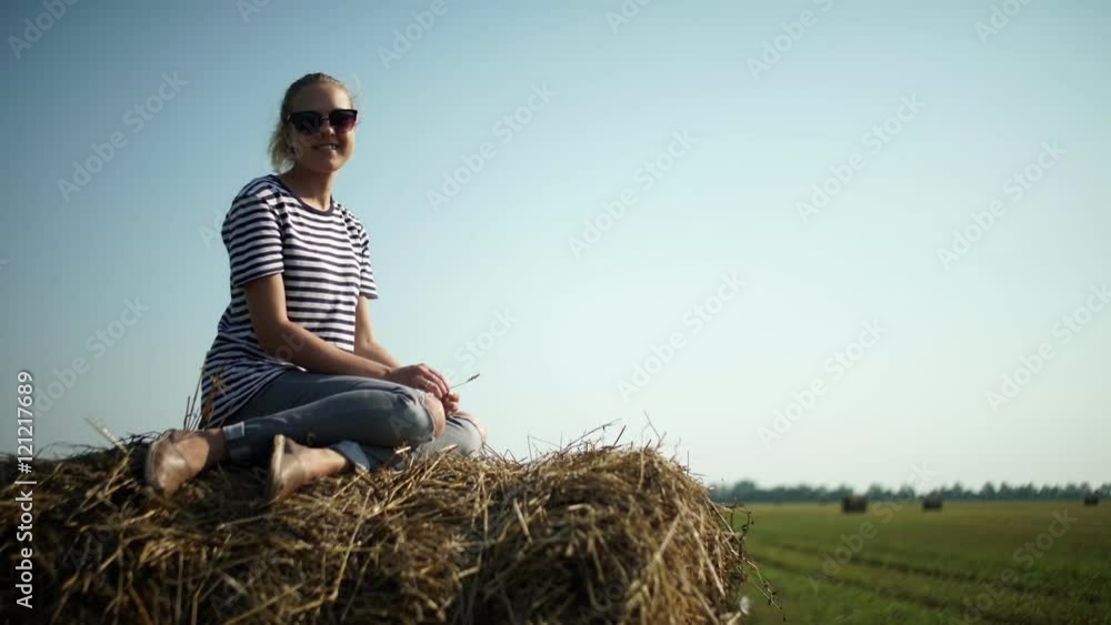 Young stylish girl in a striped t-shirt sitting on a haystack. Beautiful young girl in the field. Girl in sunglasses looks into the distance. Girl in the countryside. Pretty girl is sitting on Young stylish girl in a striped t-shirt sitting on a haystack. Beautiful young girl in the field. Girl in sunglasses looks into the distance. Girl in the countryside. Pretty girl is sitting on