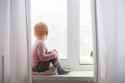 The red-haired child is sitting on window and looking at the snow