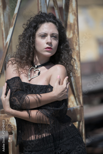 Fashion lady with curly hairs wear a crop top gauzy short sleeve shirt and shorts pose outdoor near by a rusty iron construction