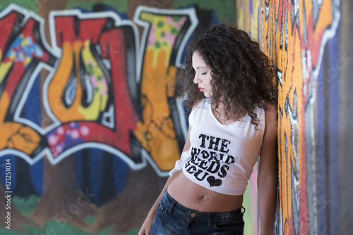 Pretty lady with curly hair wear a crop top shirt and shorts leaning on a graffiti wall